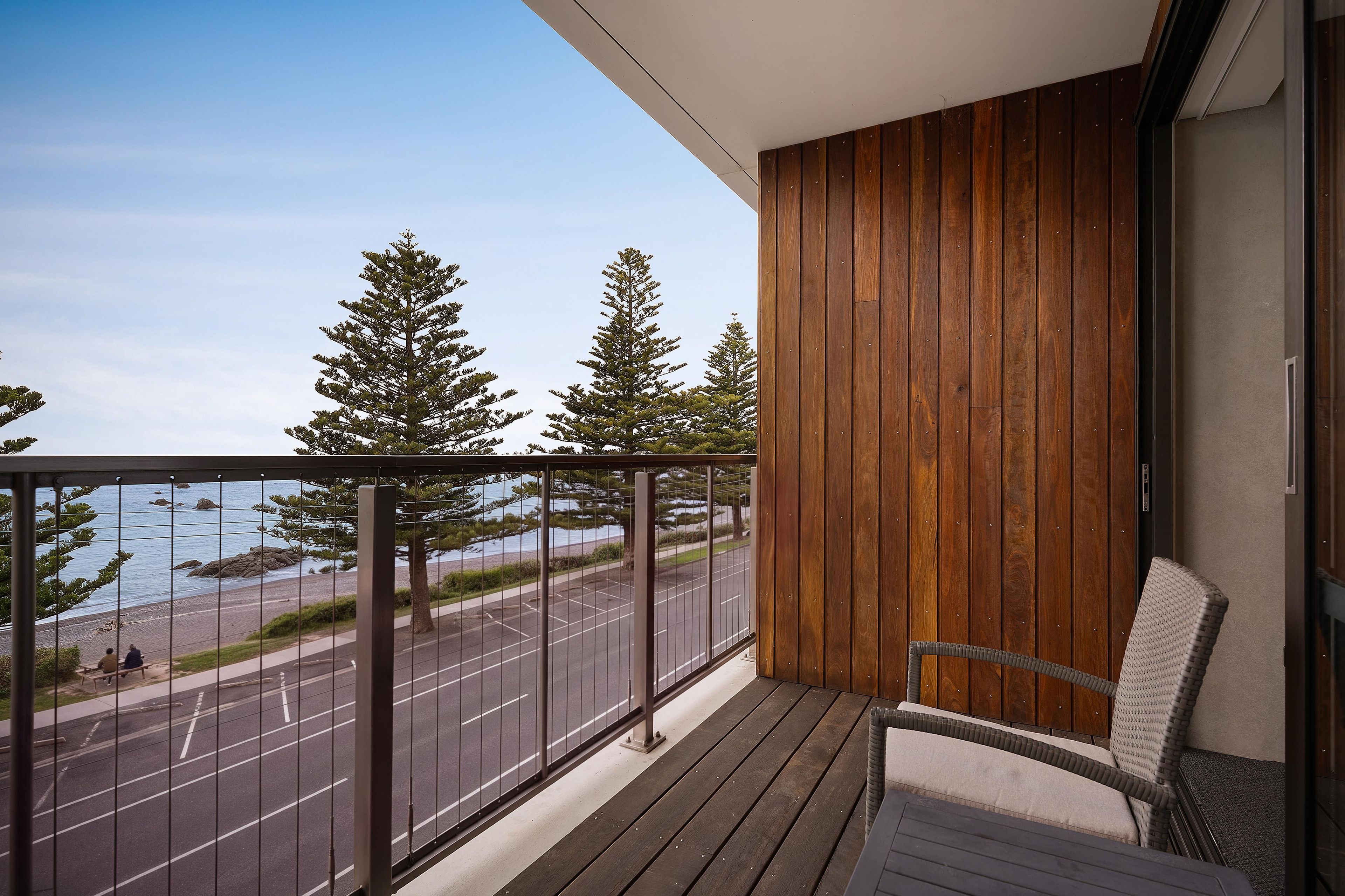 A balcony with a view of a road and trees, wooden paneling, and a wicker chair.