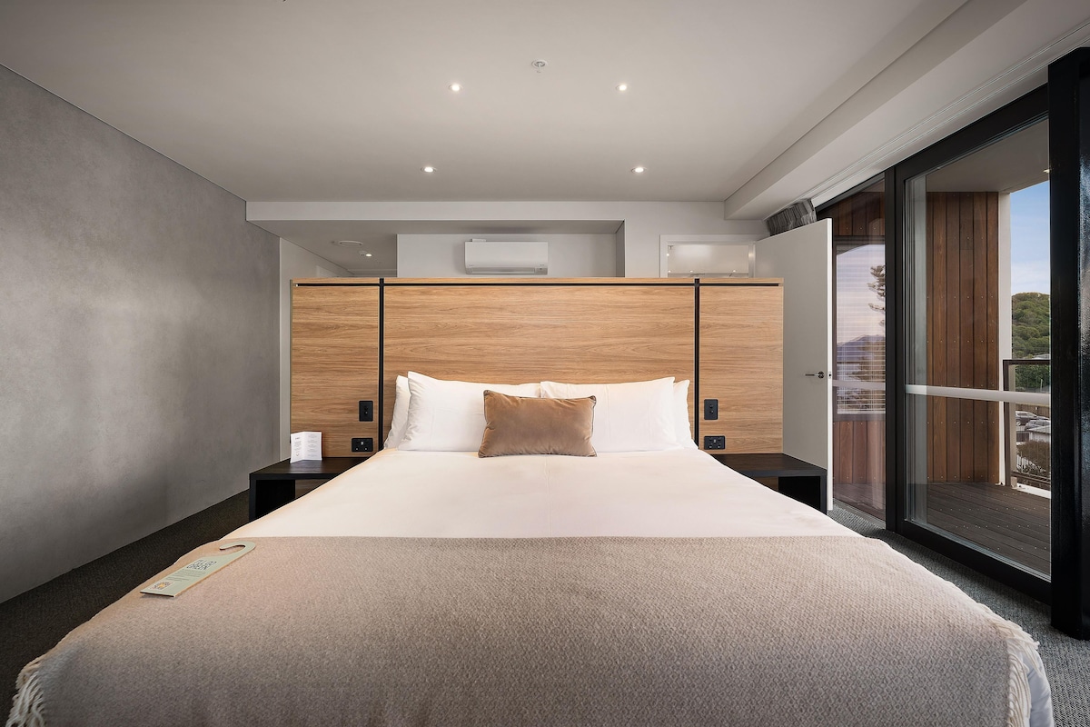 A modern hotel room with a large bed, wooden headboard, and a sliding glass door to the outside.