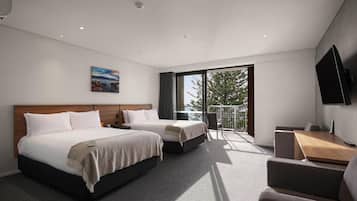 A hotel room with two beds, a desk, a chair, a TV, and a balcony with a view of trees.