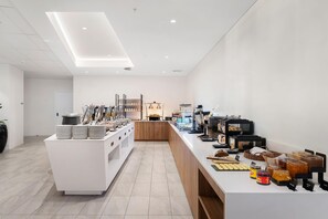 Interior moderno de una cafetería con una barra larga, máquinas de café y una variedad de alimentos a la vista.