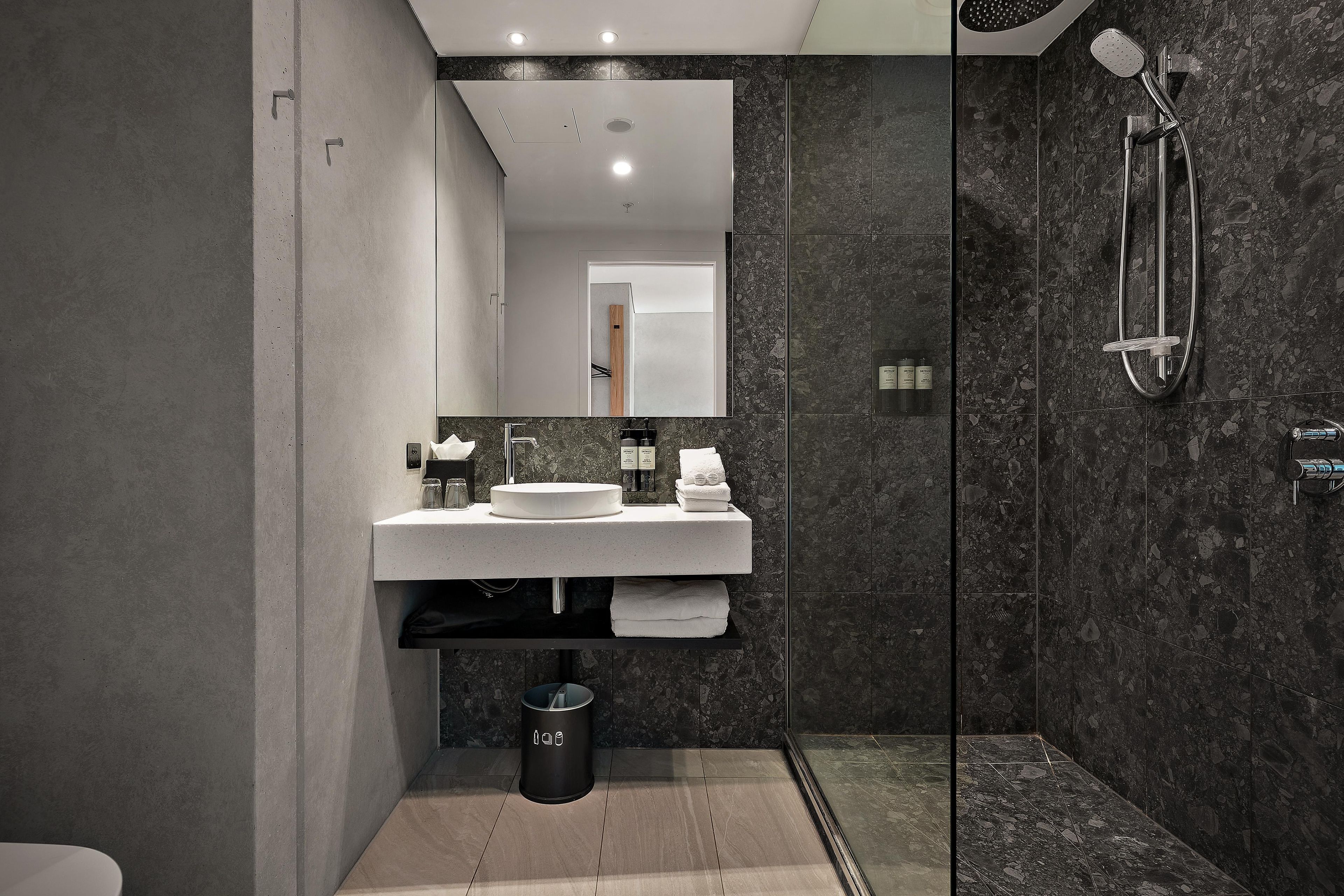 A modern bathroom with a large mirror, a round sink, a glass shower enclosure, and a trash bin.