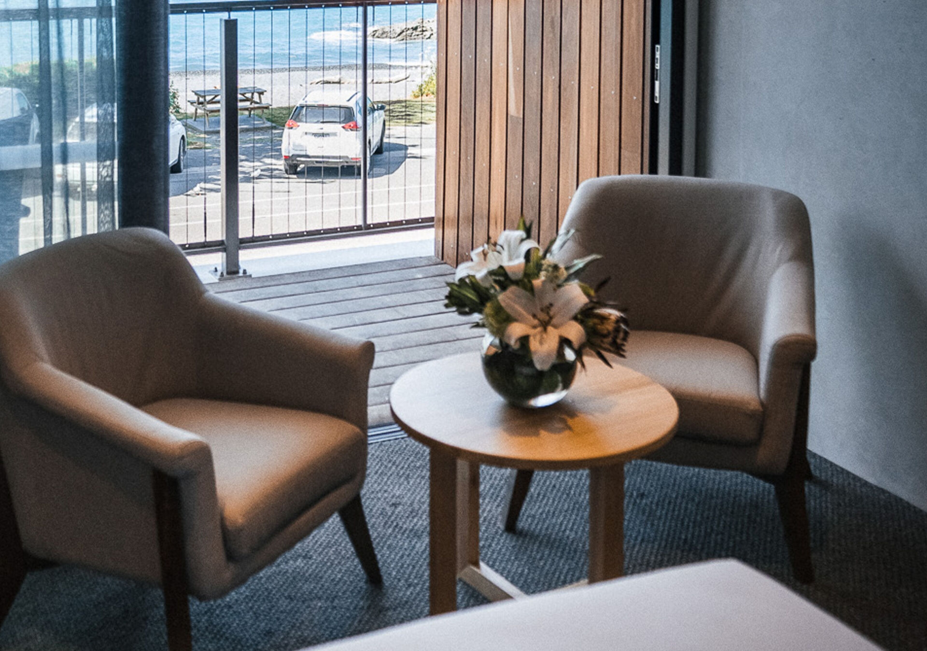 A room with two beige armchairs, a small wooden table with a flower vase, and a view of the beach and parked cars through large windows.