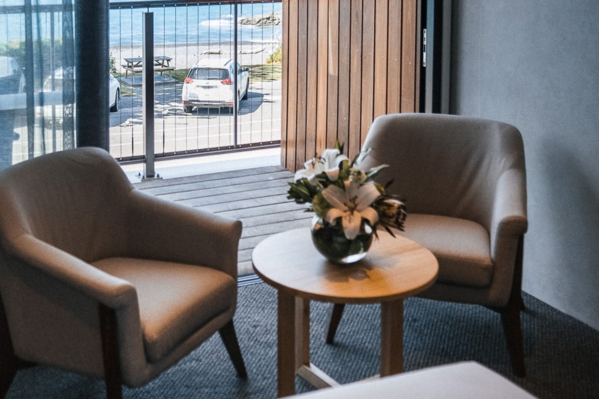 Una habitación con dos sillones beige, una mesita de madera con un florero y vistas a la playa y coches aparcados a través de amplios ventanales.