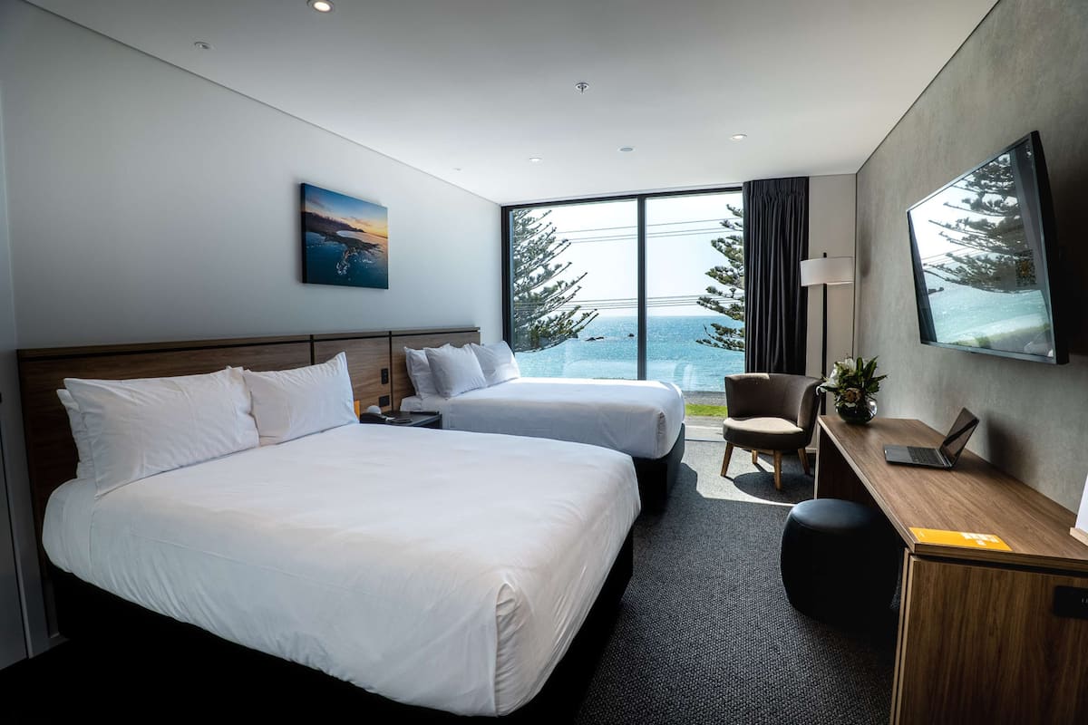 Een hotelkamer met twee bedden, een bureau, een stoel, een televisie en een groot raam met uitzicht op zee en bomen.
