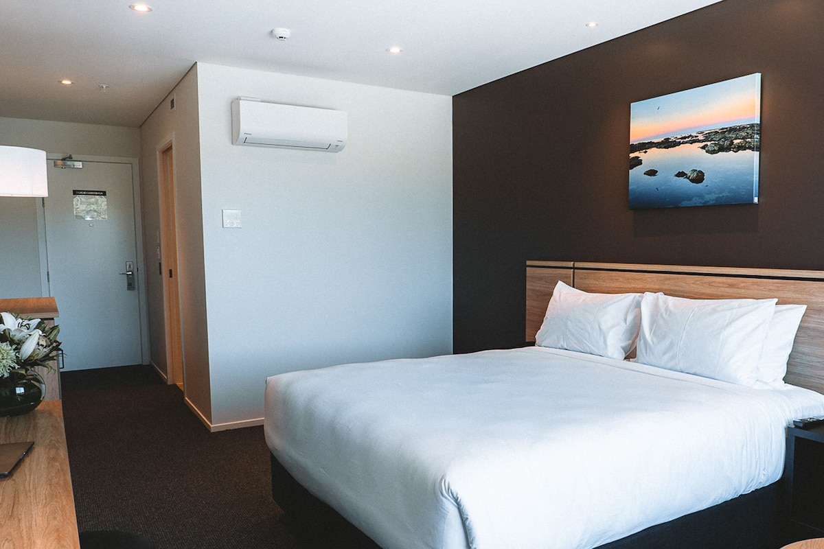 Una habitación de hotel con una cama grande, una mesita de noche, aire acondicionado instalado en la pared y un cuadro de un paisaje.