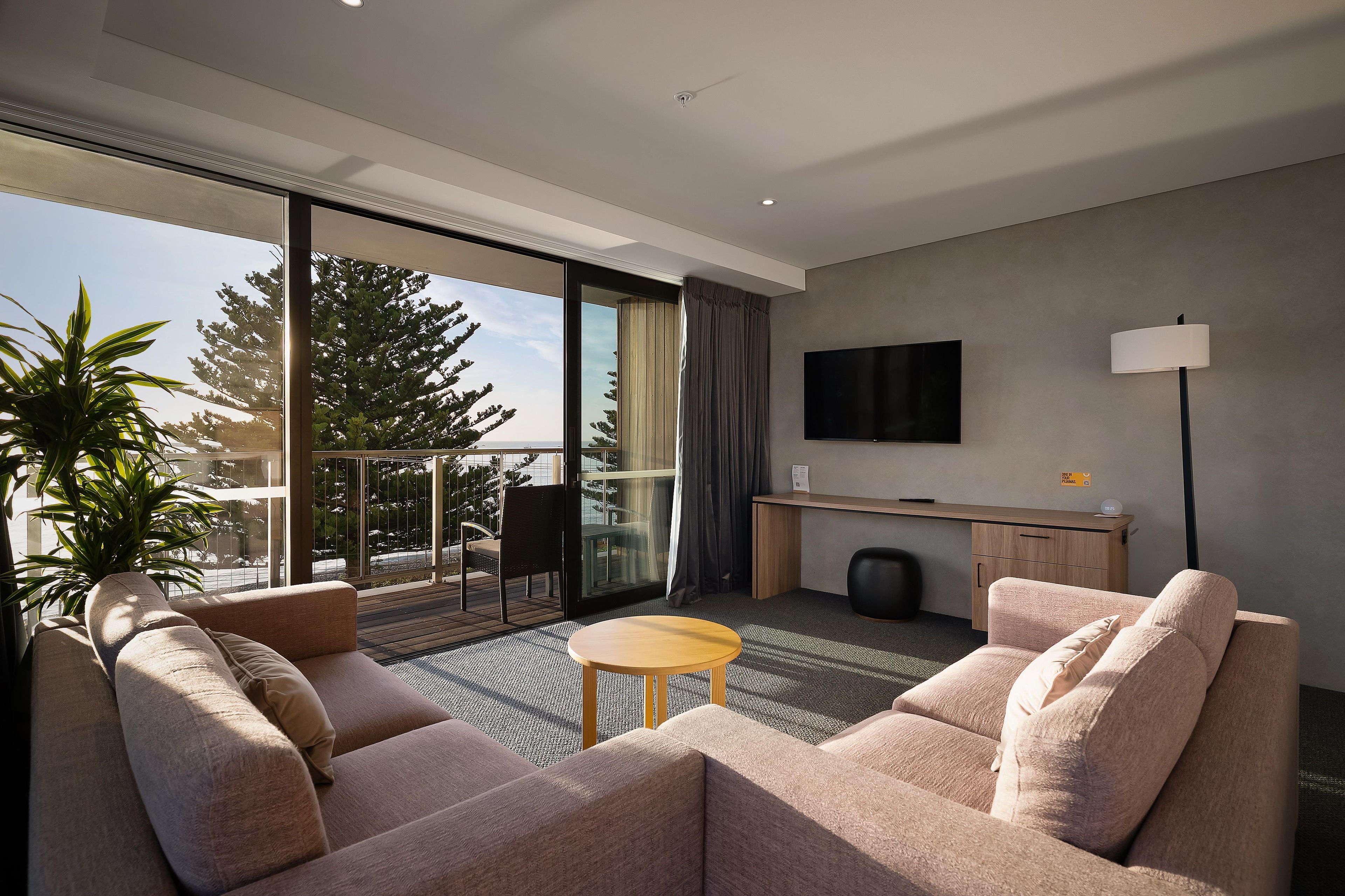 Habitación de hotel moderna con sofá, una pequeña mesa, televisor montado en la pared y balcón con vistas a árboles y una masa de agua.
