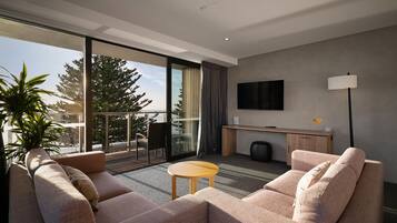 Habitación de hotel moderna con sofá, una pequeña mesa, televisor montado en la pared y balcón con vistas a árboles y una masa de agua.