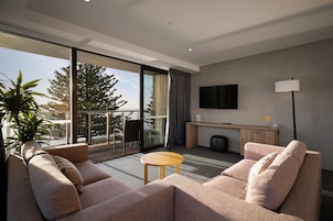 A modern hotel room with a sofa set, a small table, a TV mounted on the wall, and a balcony with a view of trees and a body of water.