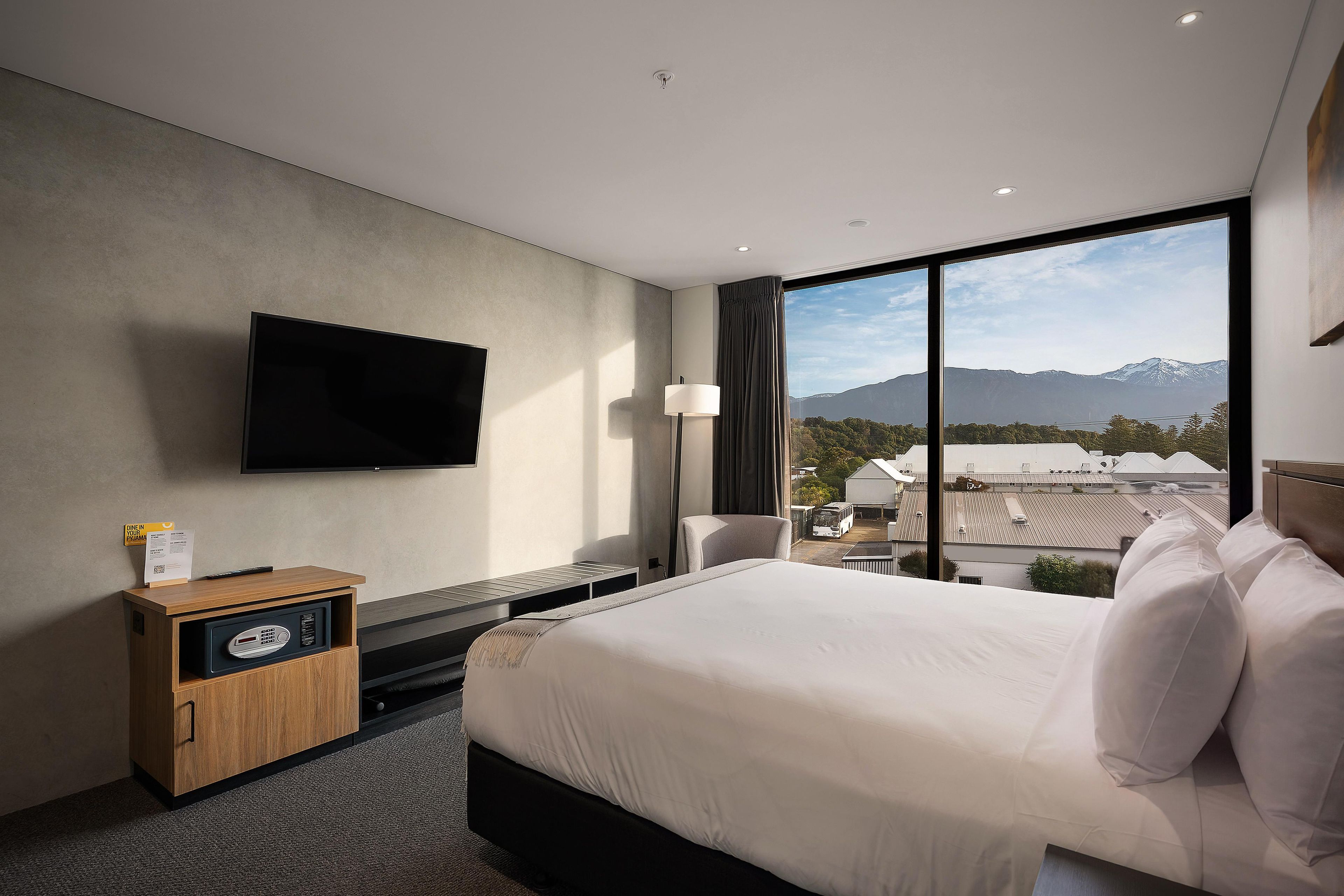 Habitación de hotel con una cama grande, un televisor de pantalla plana montado en la pared, un mueble de madera con una caja registradora, y vistas a montañas a través de un ventanal amplio.