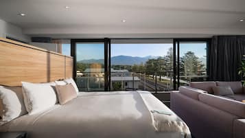 Ein modernes Hotelzimmer mit einem großen Bett, einer Couch und Blick auf eine Berglandschaft durch raumhohe Fenster.