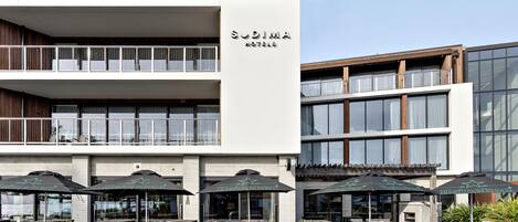 A modern hotel with a two-story structure, outdoor seating with umbrellas, and a clear sky.