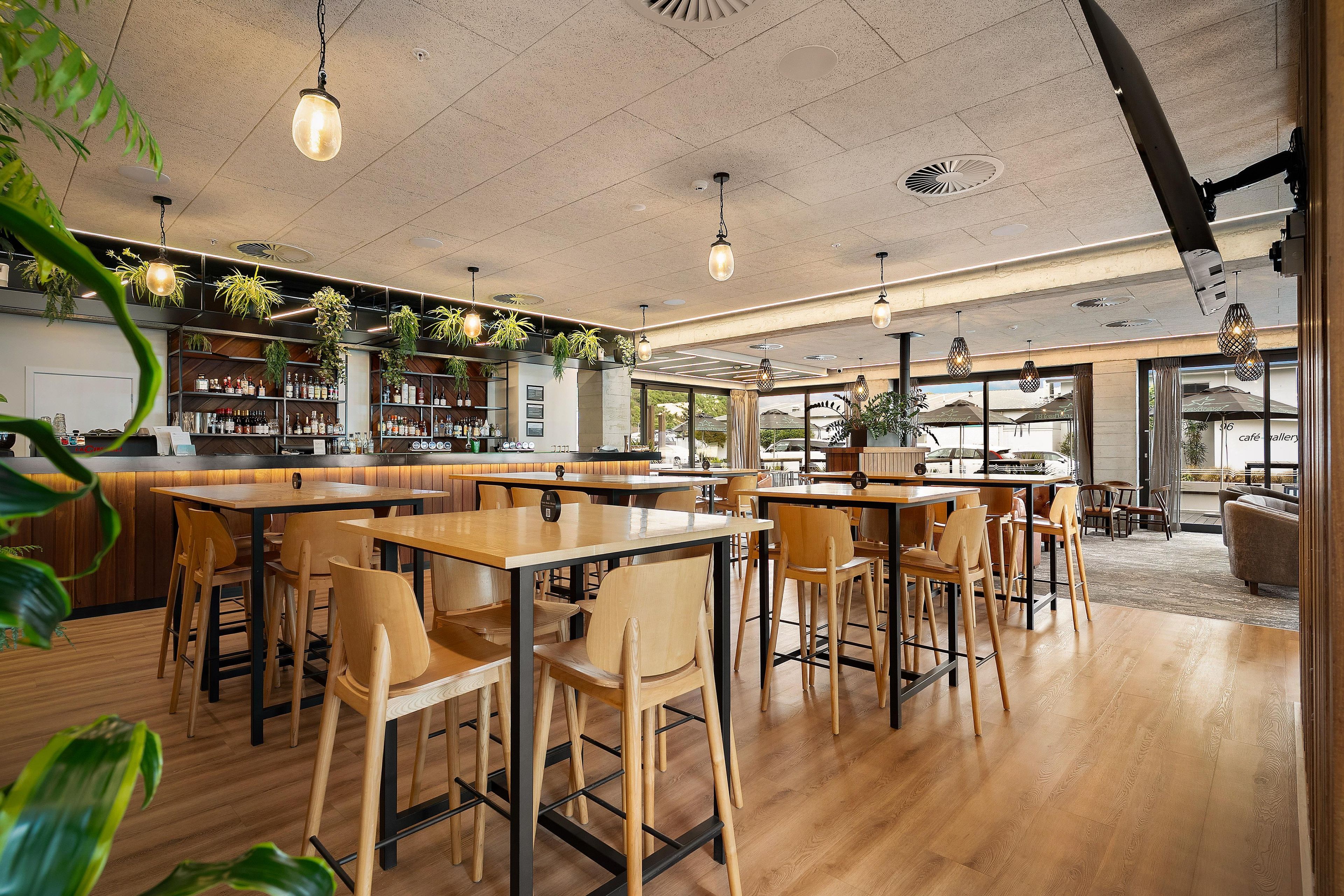 Ein geräumiges Café mit Holztischen und -stühlen, einer Bar mit Flaschenregalen und hängenden Pflanzen.