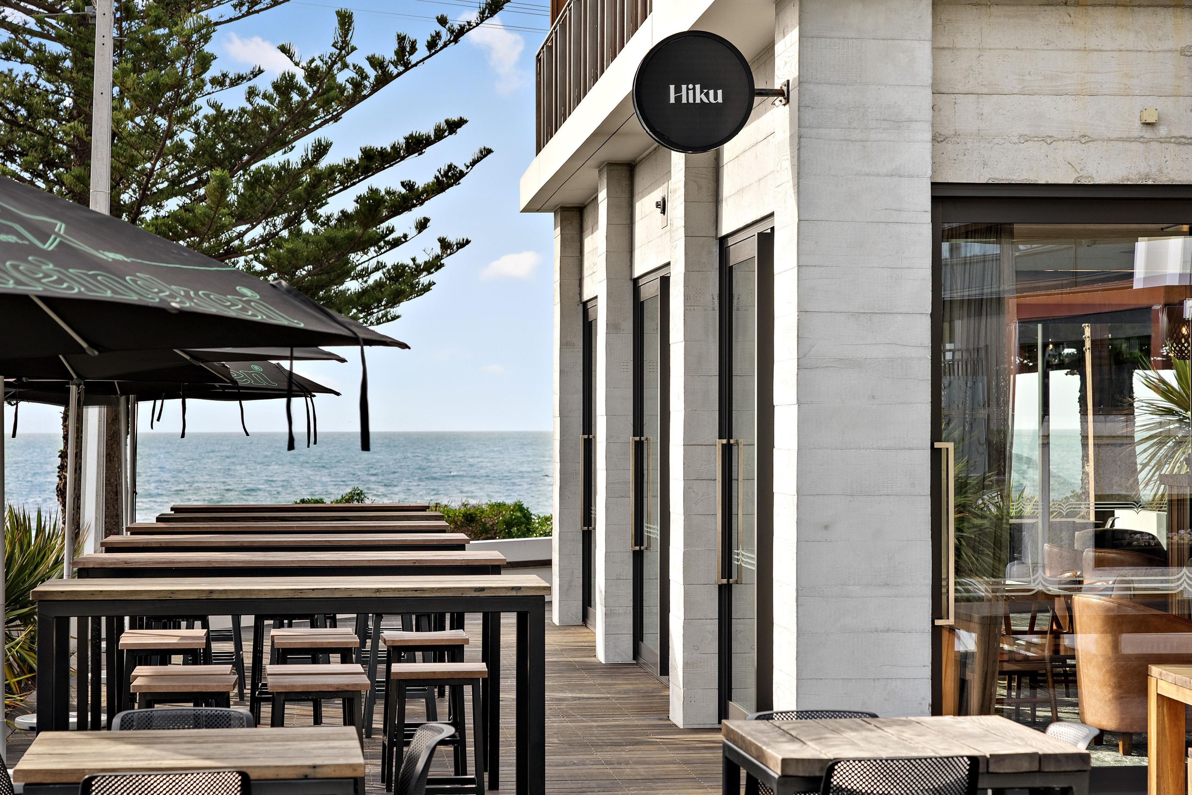 An outdoor seating area with a view of the sea, a sign reading 'Hiku', and a modern building facade.