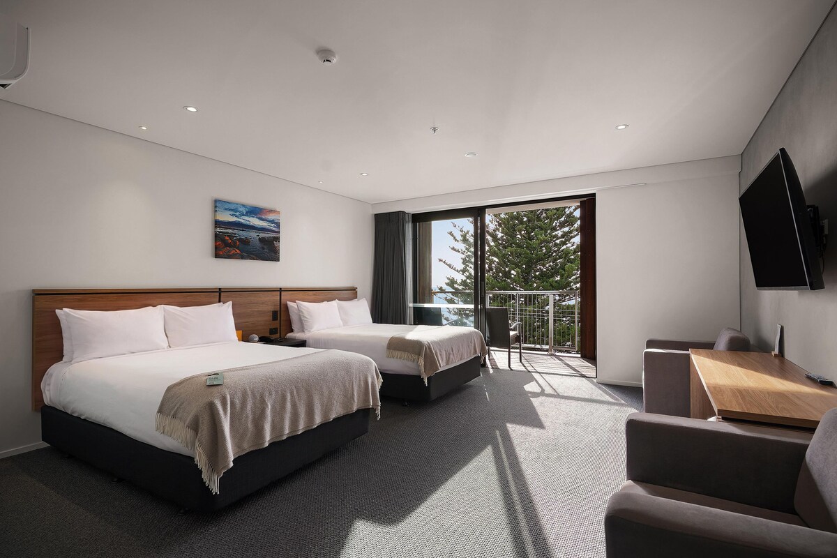Habitación de hotel con dos camas, un escritorio, una silla, un televisor y un balcón con vistas a árboles.