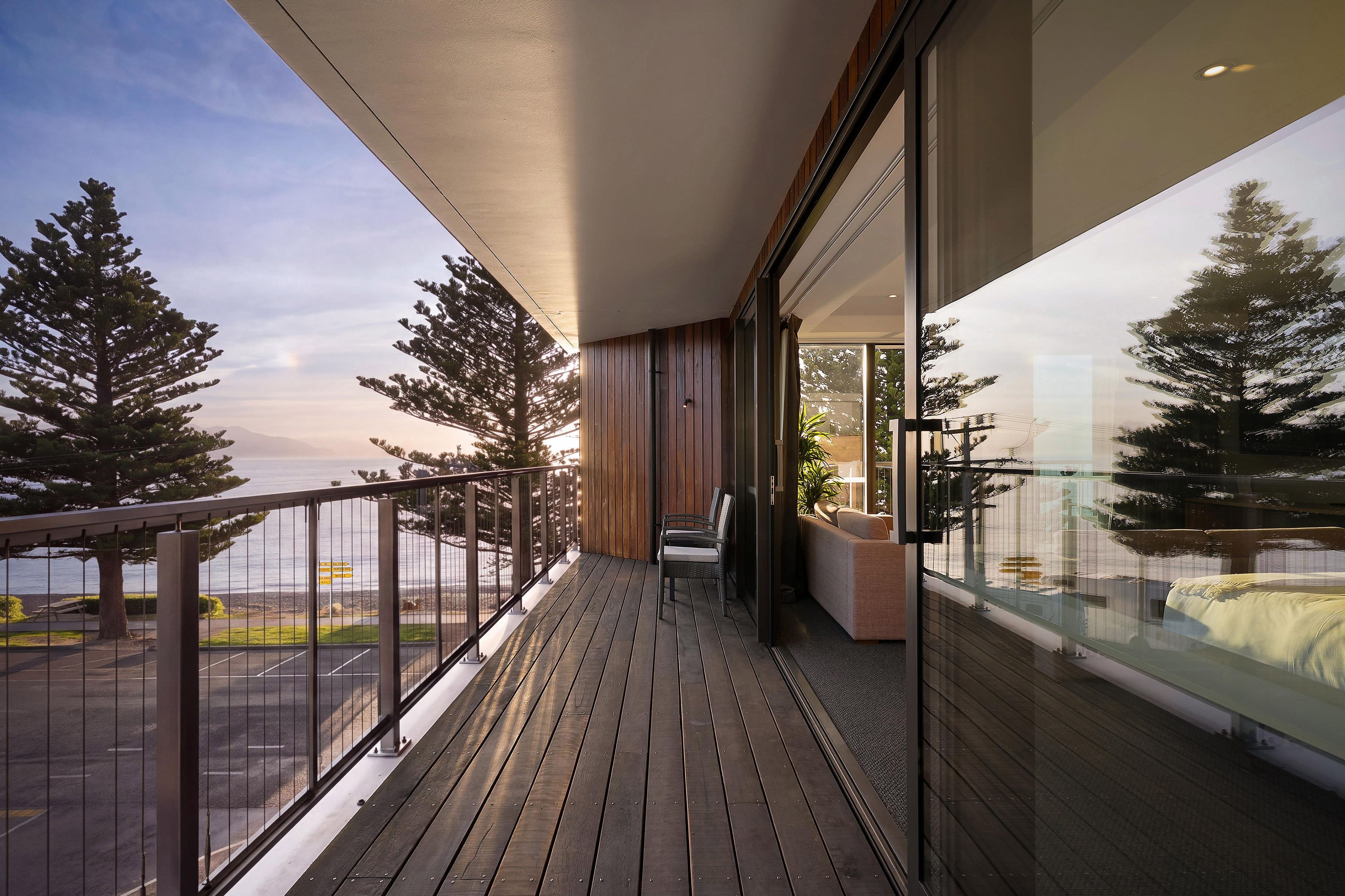 A balcony with a view of the sea and trees, wooden flooring, and a seating area with a sofa and chairs.