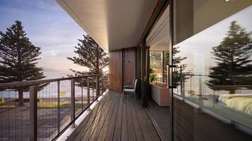 A balcony with a view of the sea and trees, wooden flooring, and a seating area with a sofa and chairs.