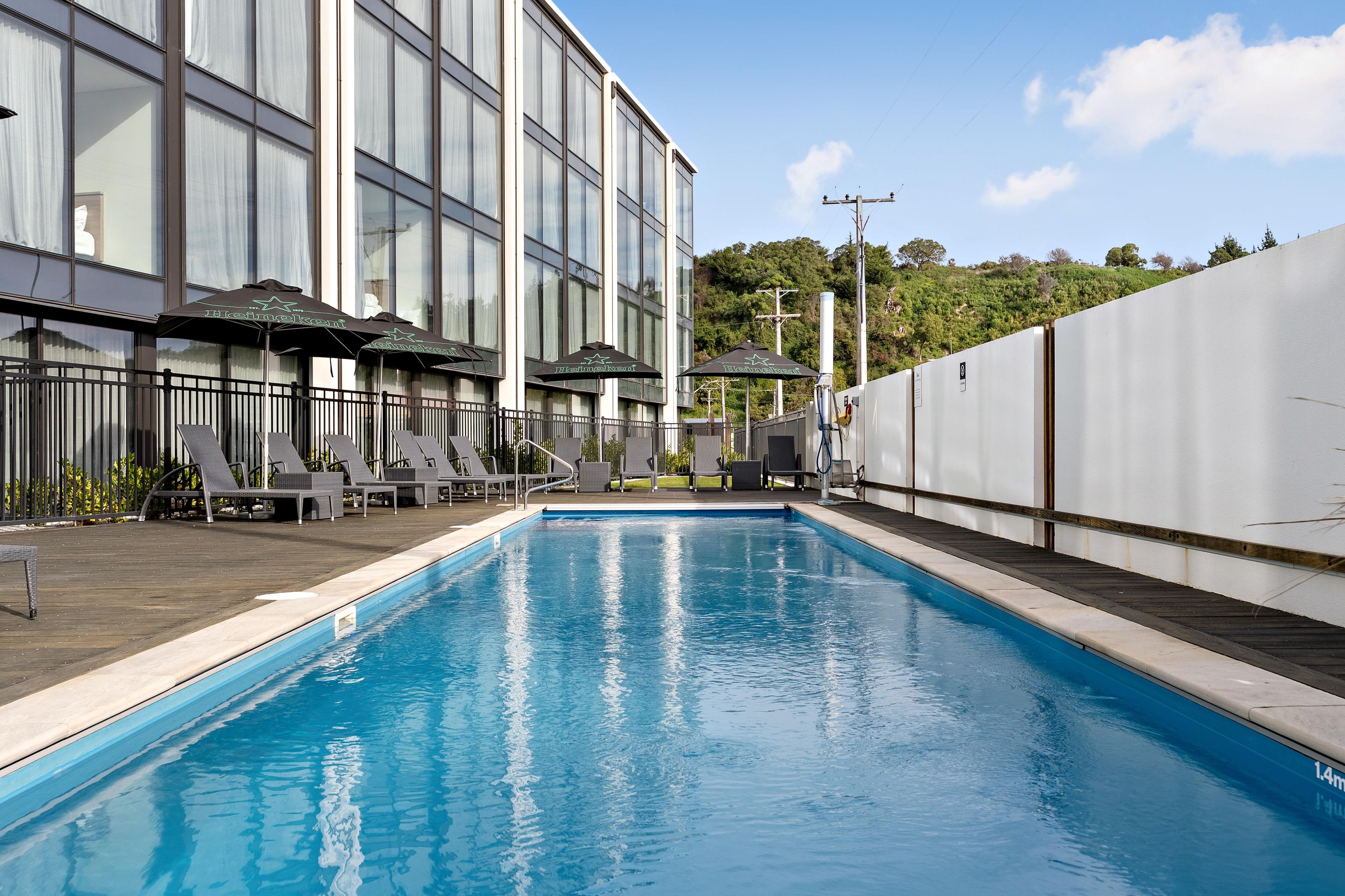 A swimming pool with lounge chairs and umbrellas, a building with large windows, and a view of a hilly landscape.