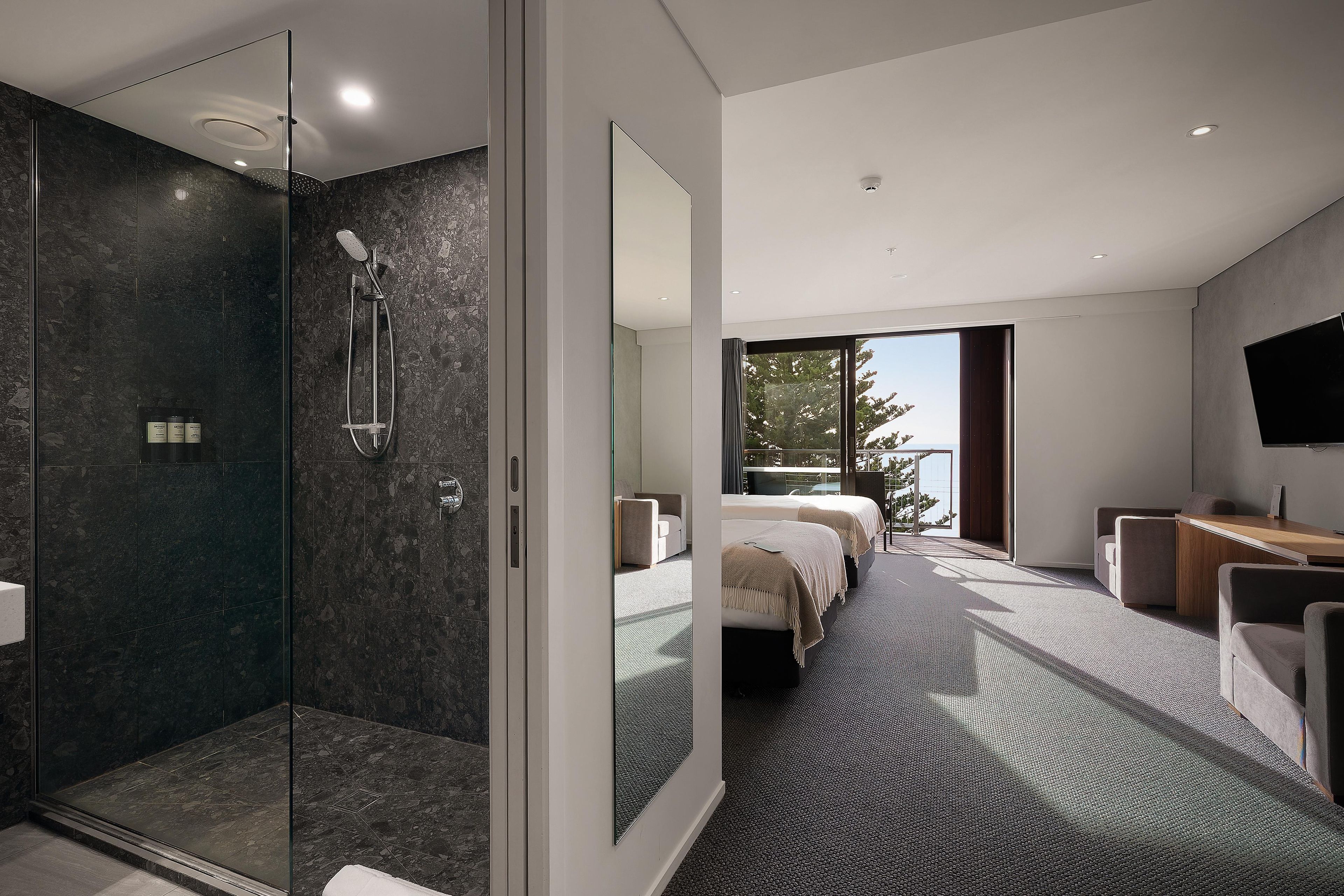 A modern hotel room with a glass-enclosed shower, a large bed, a sitting area, and a view of the outdoors.