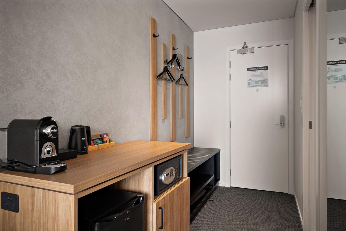 Habitación de hotel con un escritorio de madera, una cafetera, un altavoz negro y una puerta con un plan de evacuación de emergencia.