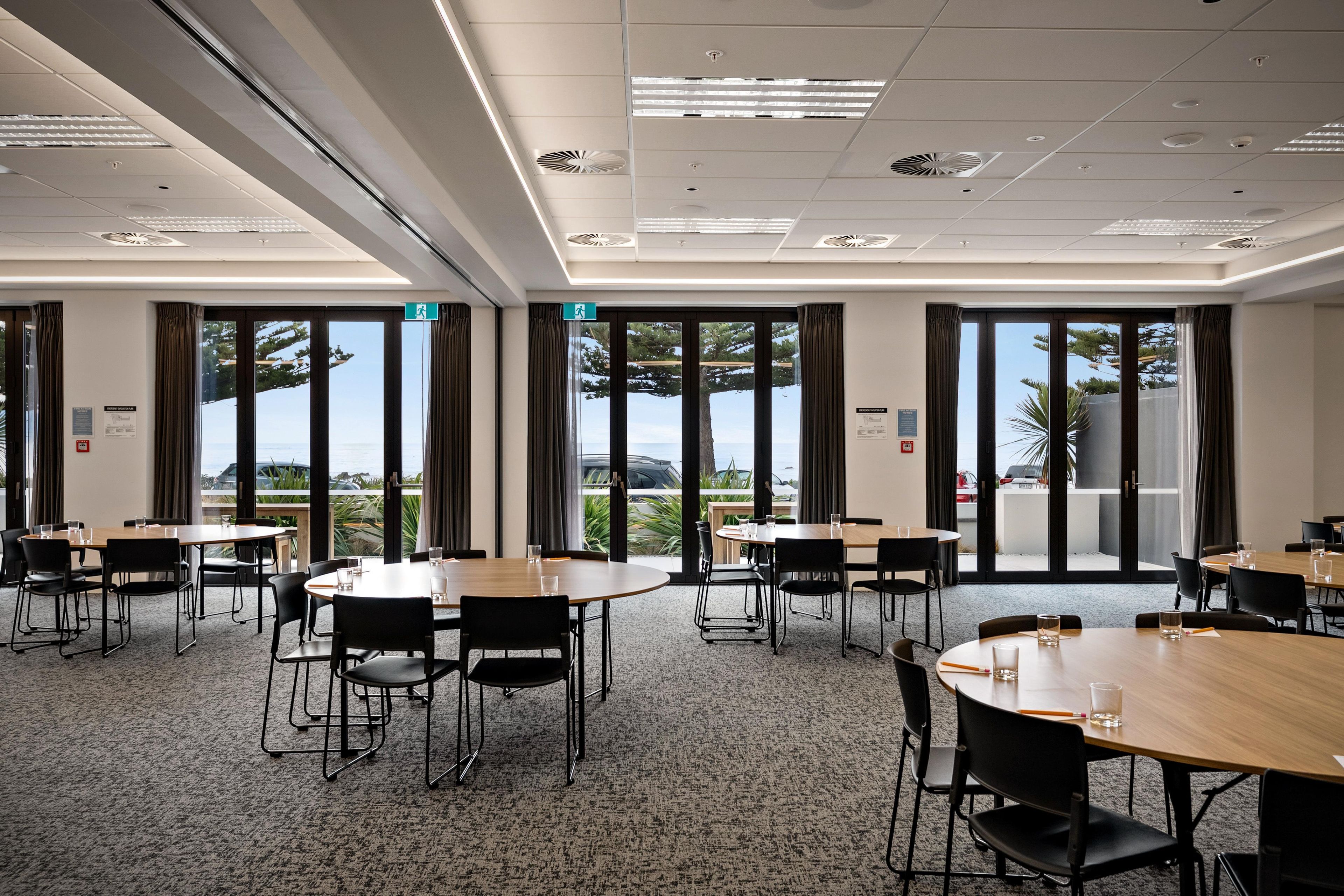 A conference room with multiple tables and chairs, large windows with curtains, and a view of the outdoors.
