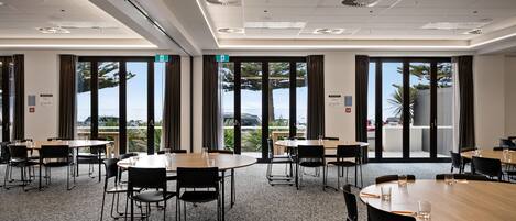 A conference room with multiple tables and chairs, large windows with curtains, and a view of the outdoors.