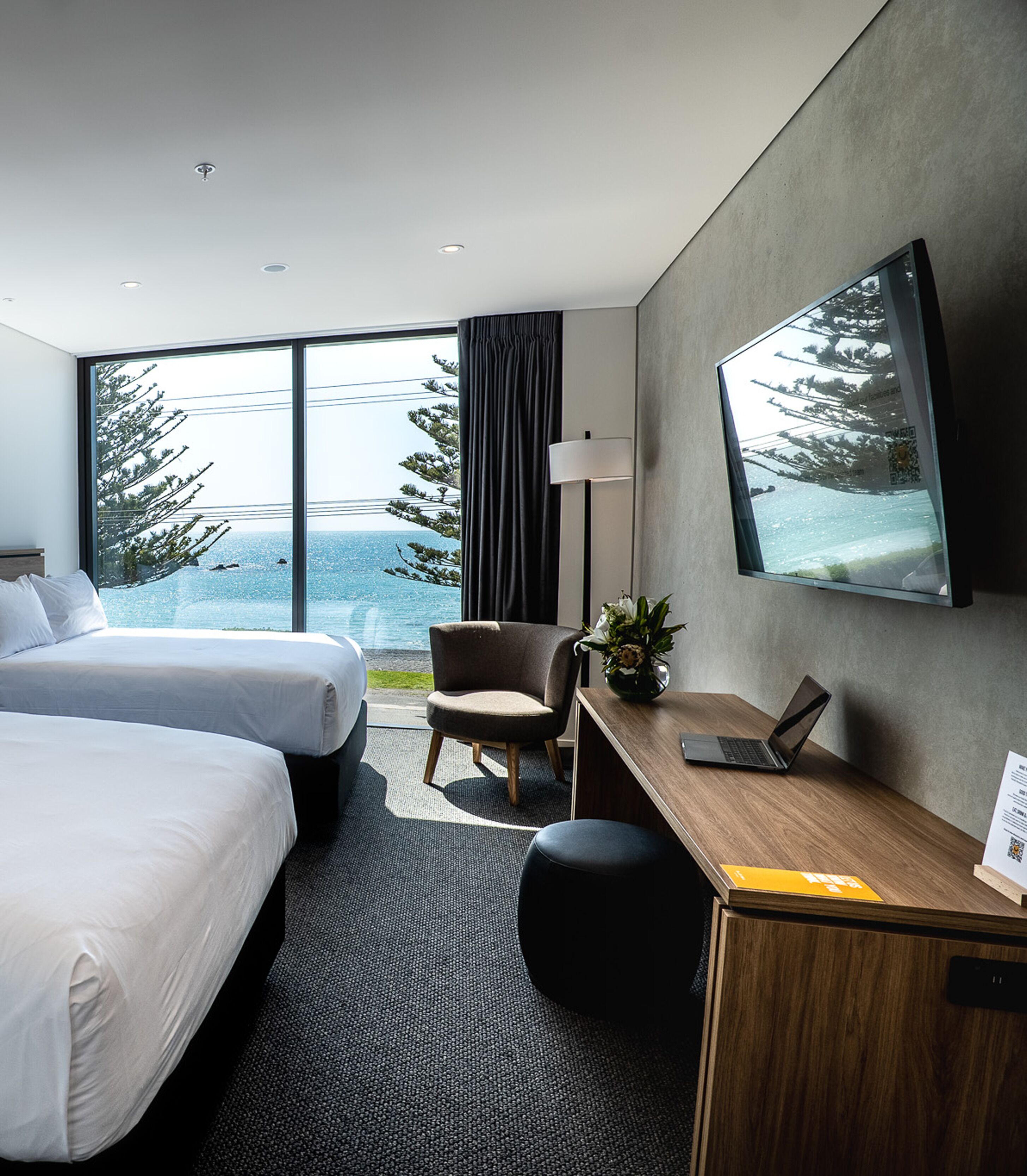 A hotel room with two beds, a desk with a laptop, a chair, a potted plant, and a large window with a view of trees and the sea.
