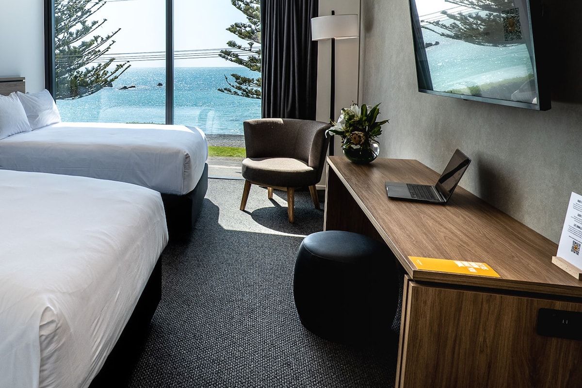Habitación de hotel con dos camas, un escritorio con una computadora portátil, una silla, una planta en maceta y un amplio ventanal con vistas a árboles y al mar.