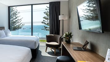 A hotel room with two beds, a desk with a laptop, a chair, a potted plant, and a large window with a view of trees and the sea.