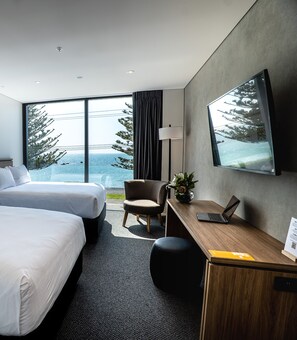 Habitación de hotel con dos camas, un escritorio con una computadora portátil, una silla, una planta en maceta y un amplio ventanal con vistas a árboles y al mar.