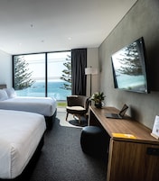 Habitación de hotel con dos camas, un escritorio con una computadora portátil, una silla, una planta en maceta y un amplio ventanal con vistas a árboles y al mar.