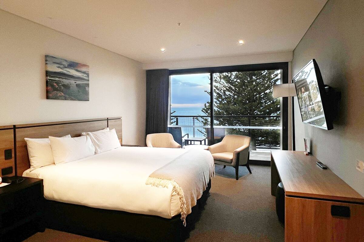 Een hotelkamer met een groot bed, een bureau, een stoel en een balkon met uitzicht op zee.