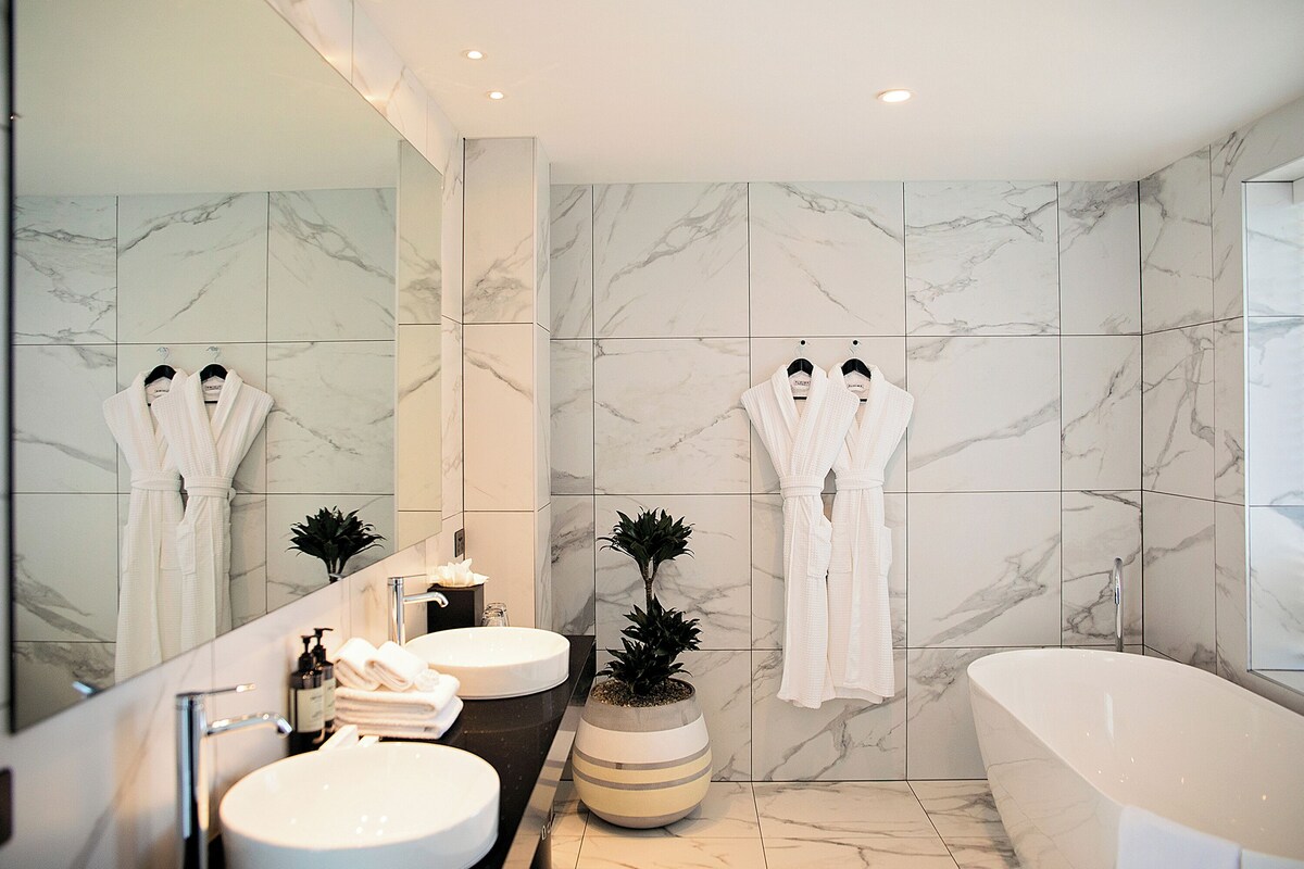 Un baño moderno con un espejo grande, dos lavamanos, una bañera y paredes de mármol.