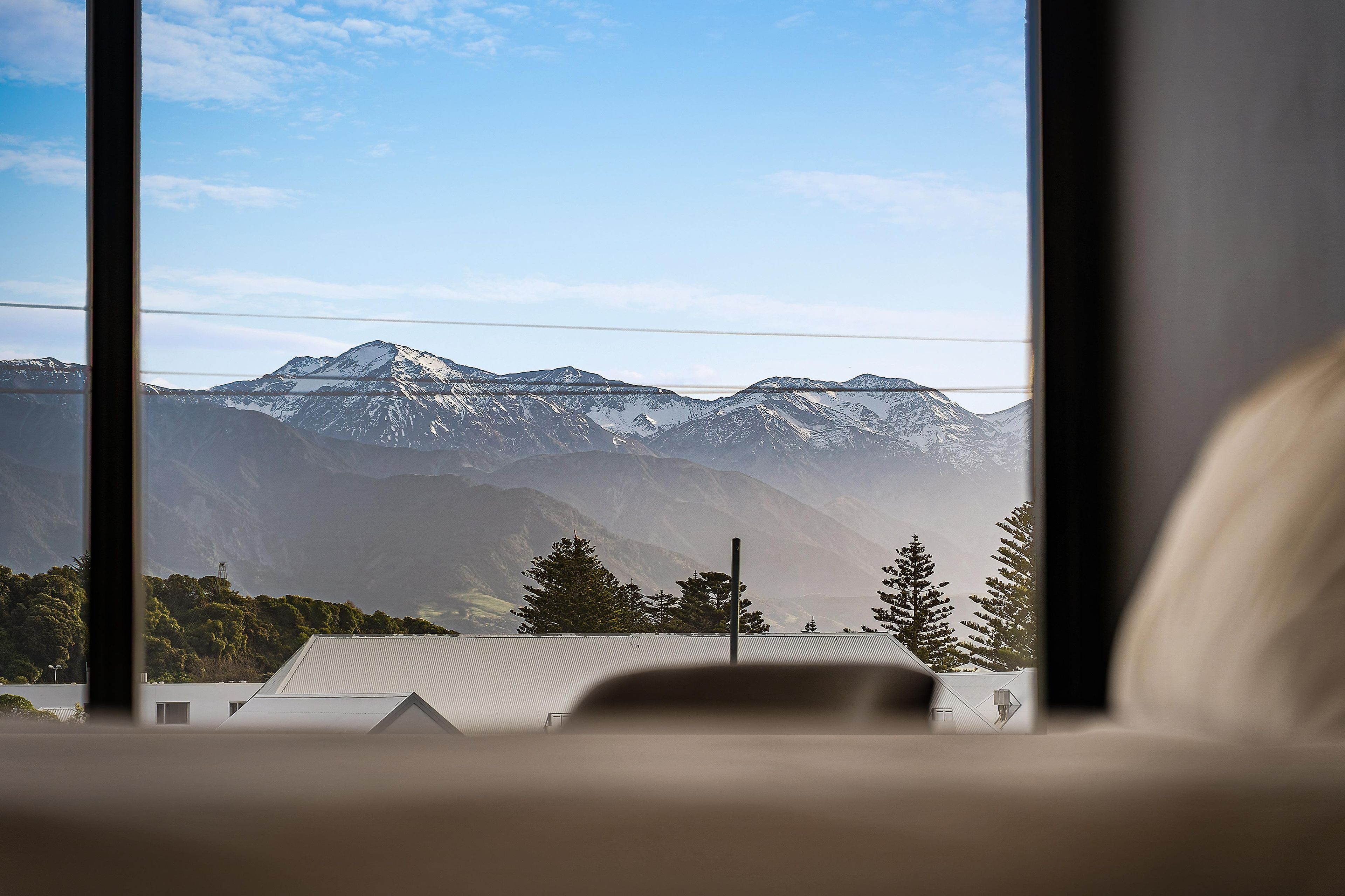 Vista de montanhas cobertas de neve através de uma janela, com um edifício e árvores ao primeiro plano.