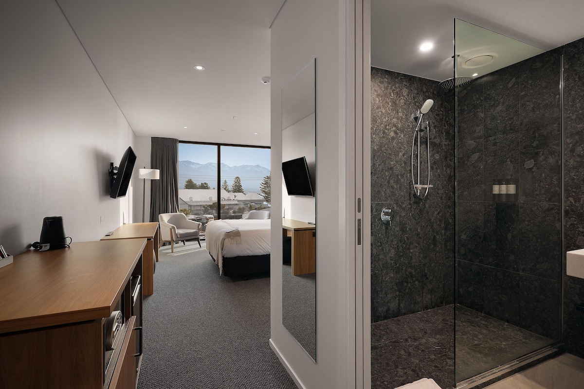A hotel room with a bed, a desk, a chair, a TV, a shower area, and a view of mountains through the window.