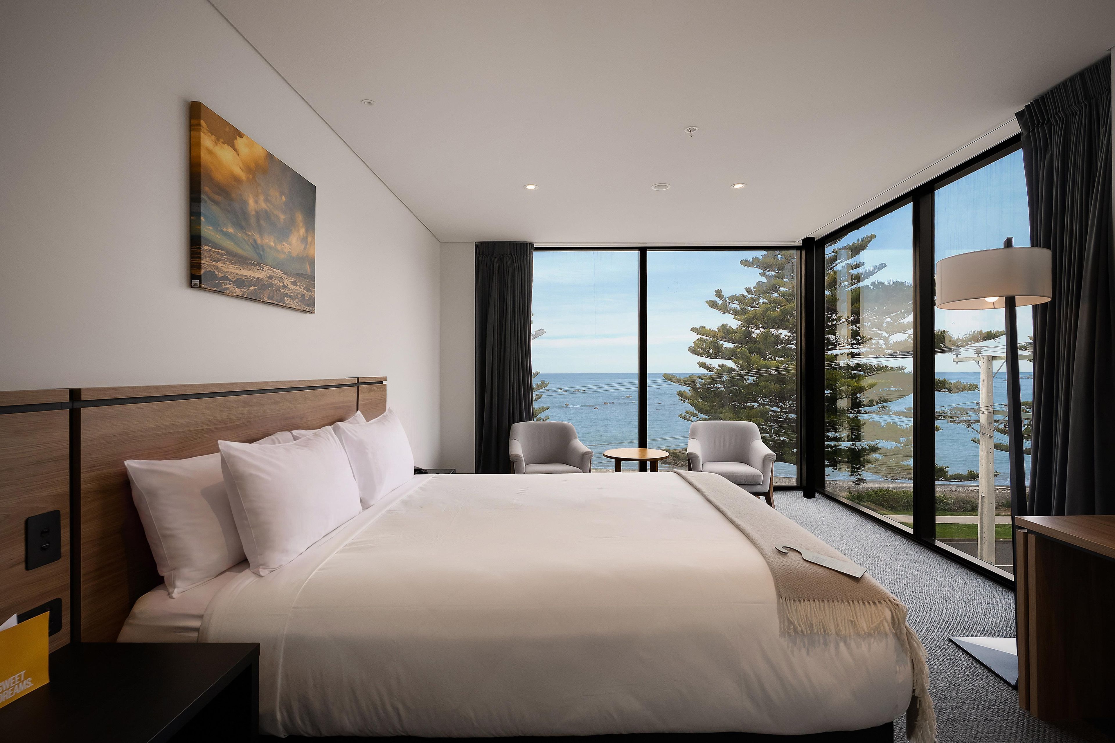 A hotel room with a large bed, a small table, two chairs, a painting on the wall, and a view of the sea through large windows.