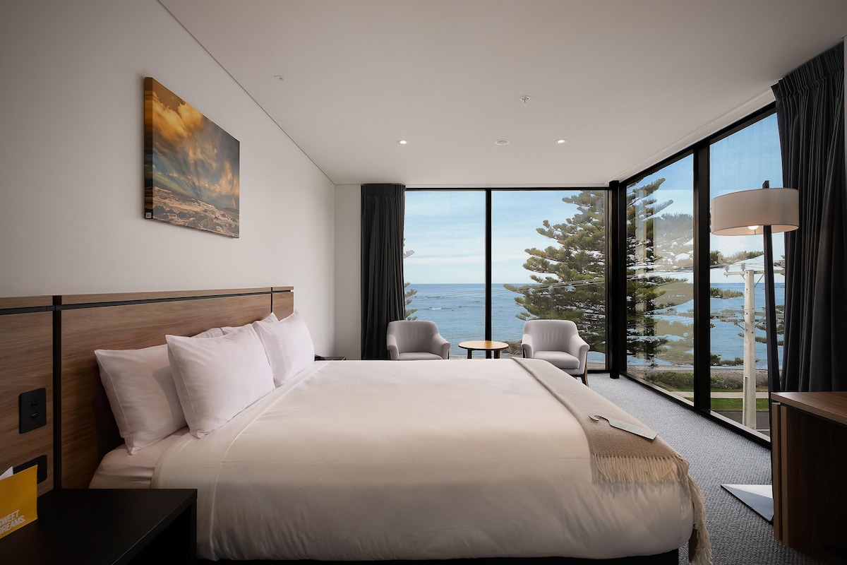 A hotel room with a large bed, a small table, two chairs, a painting on the wall, and a view of the sea through large windows.