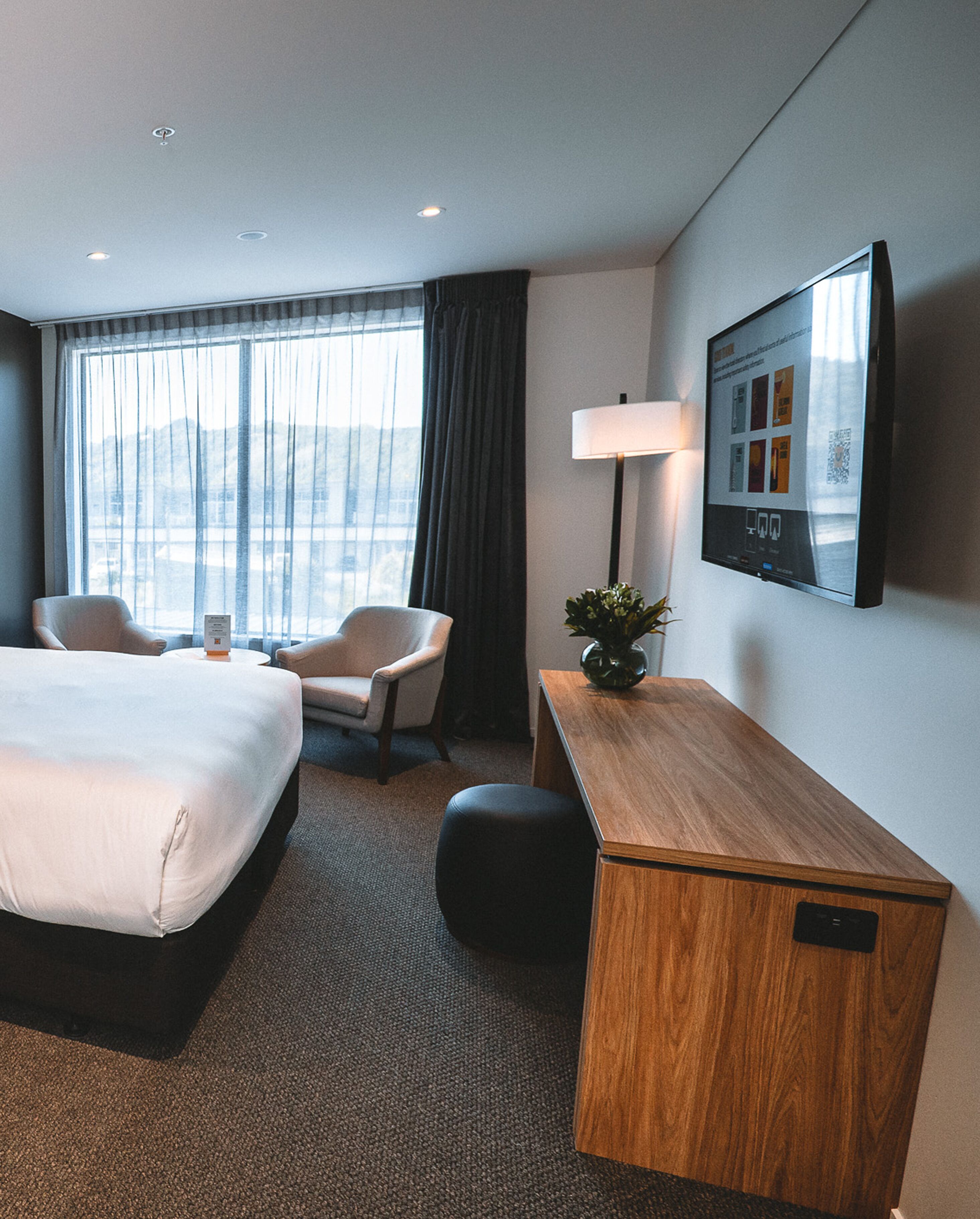 A hotel room with a bed, two chairs, a desk, a lamp, and a TV on the wall.