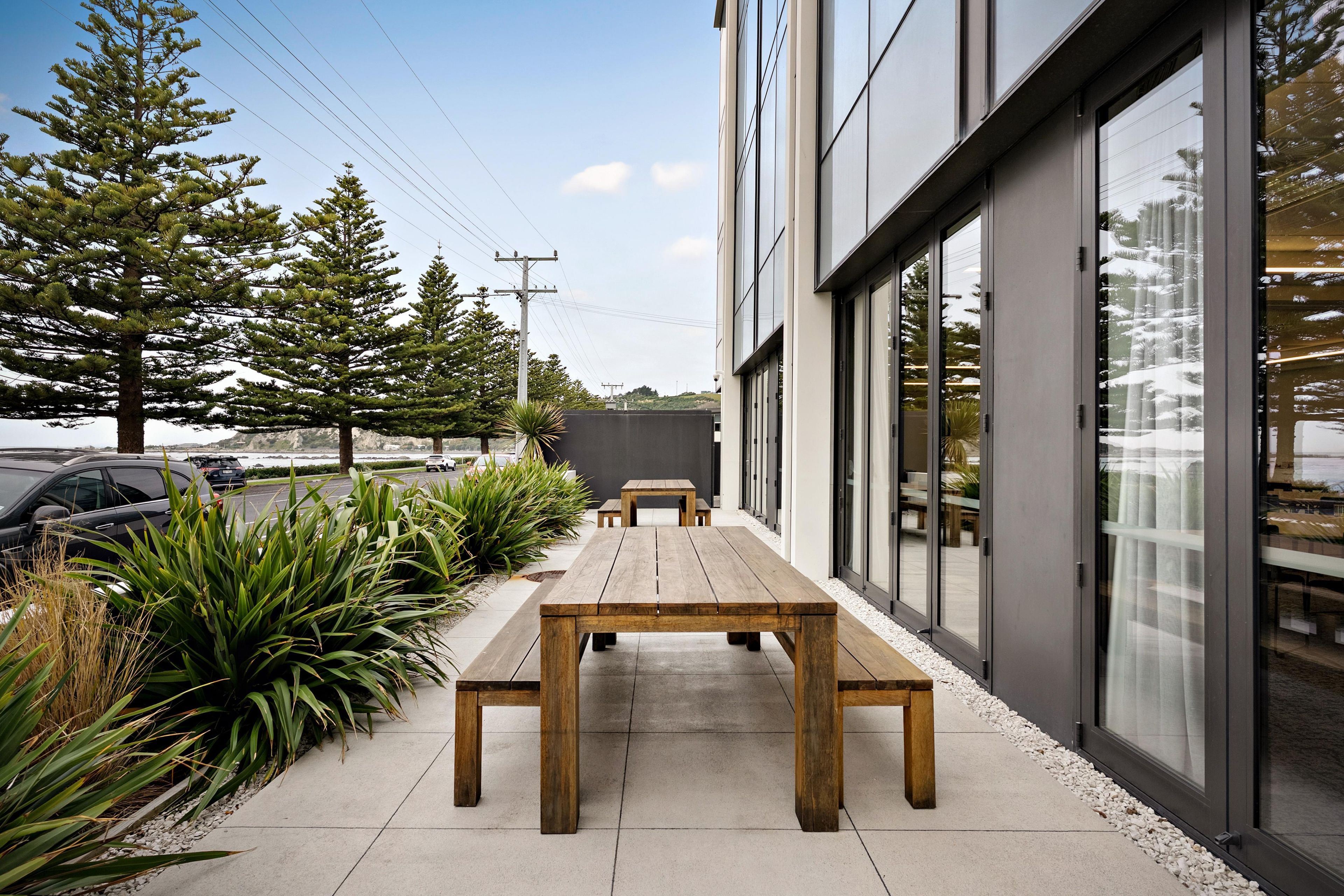 Een terras met houten tafels en banken, omgeven door planten en een gebouw met grote ramen.