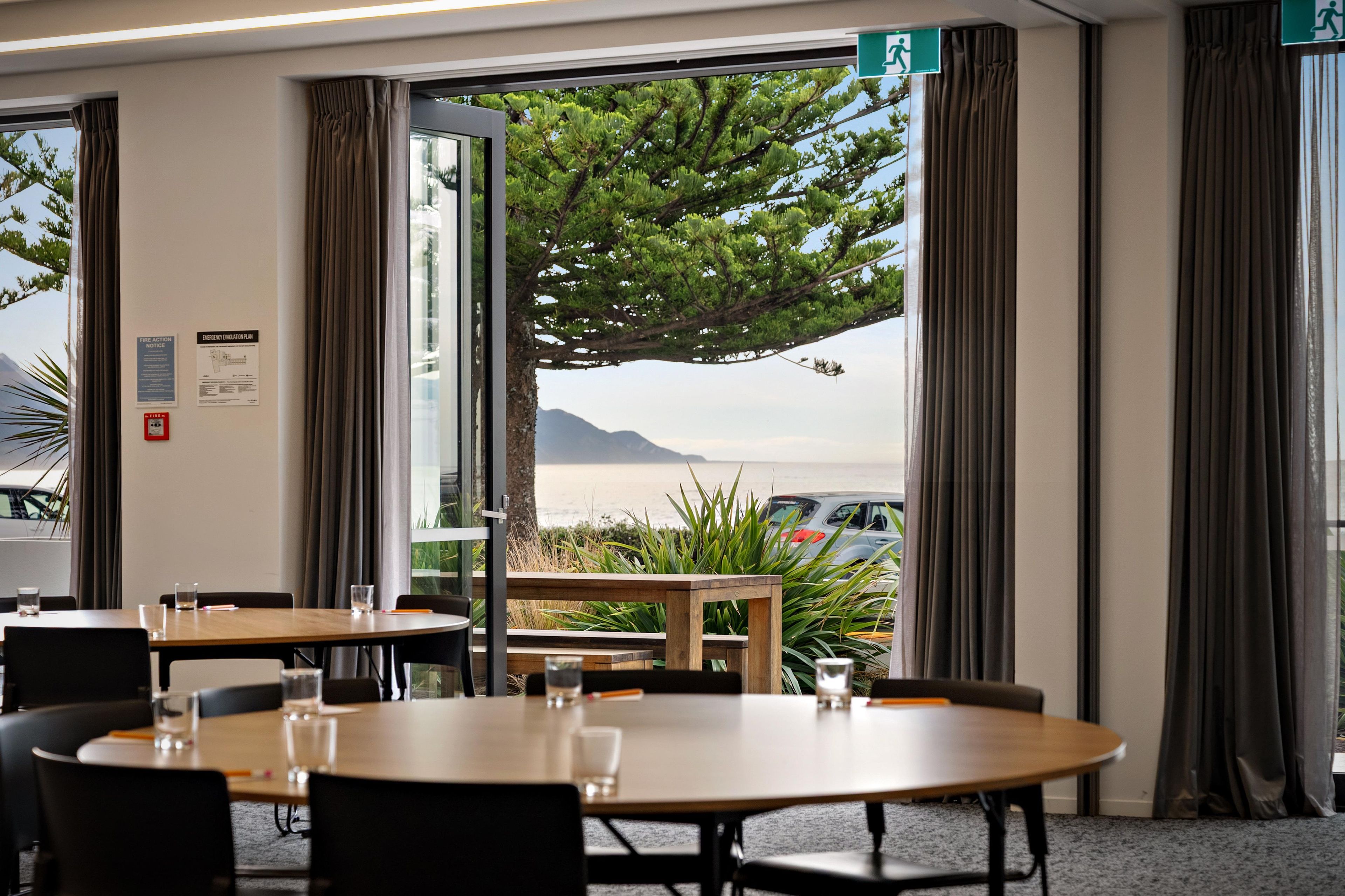 Ein Konferenzraum mit einem großen Tisch und Stühlen, Blick auf das Meer und die Berge durch die Fenster sowie Notausgangsschildern an der Wand.