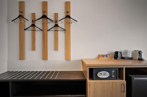 Habitación de hotel con un escritorio de madera, un refrigerador negro, una cafetera y un conjunto de estantes de madera con perchas negras.