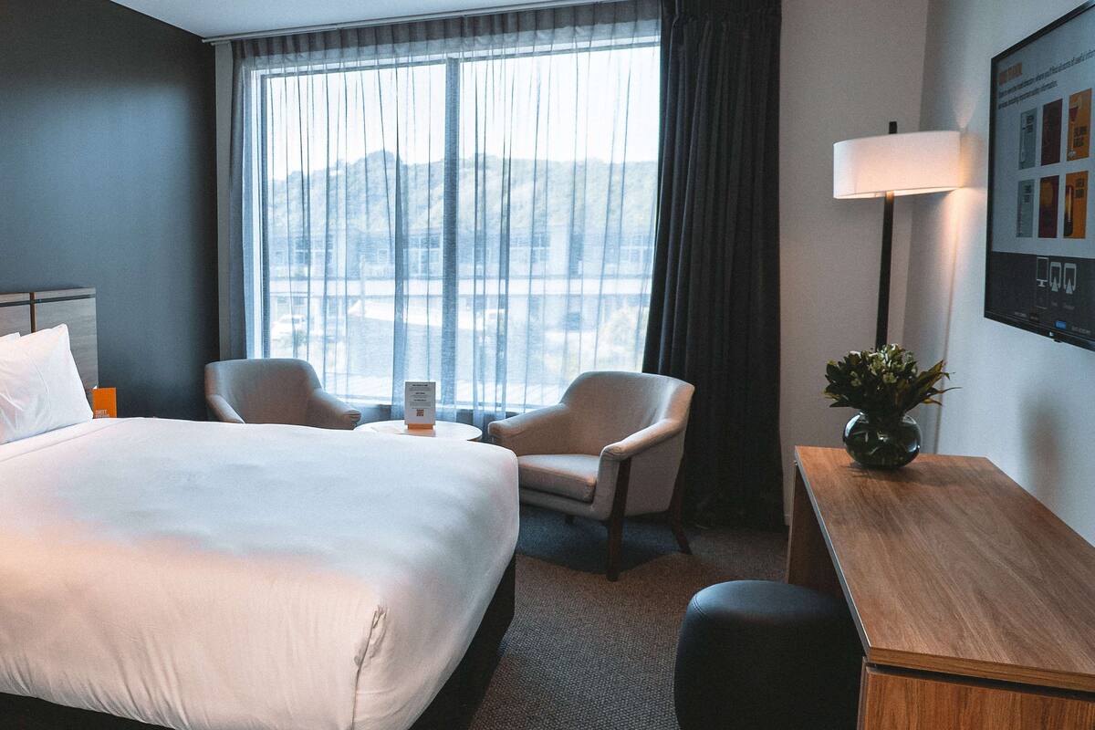 Een hotelkamer met een groot bed, twee fauteuils, een bureau, een lamp en een stadszicht door dunne gordijnen.