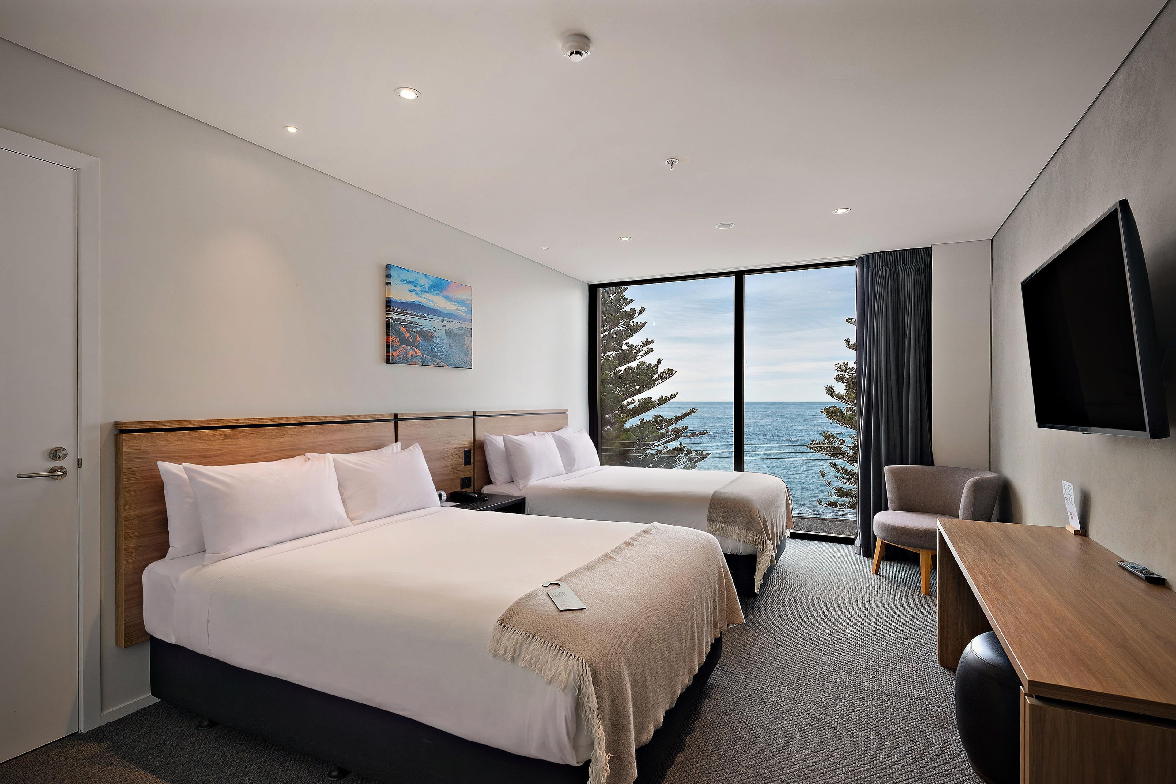 A hotel room with two beds, a desk, a TV, and a large window with a view of the sea.