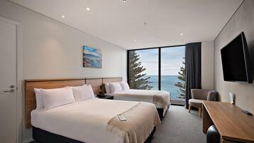 A hotel room with two beds, a desk, a TV, and a large window with a view of the sea.