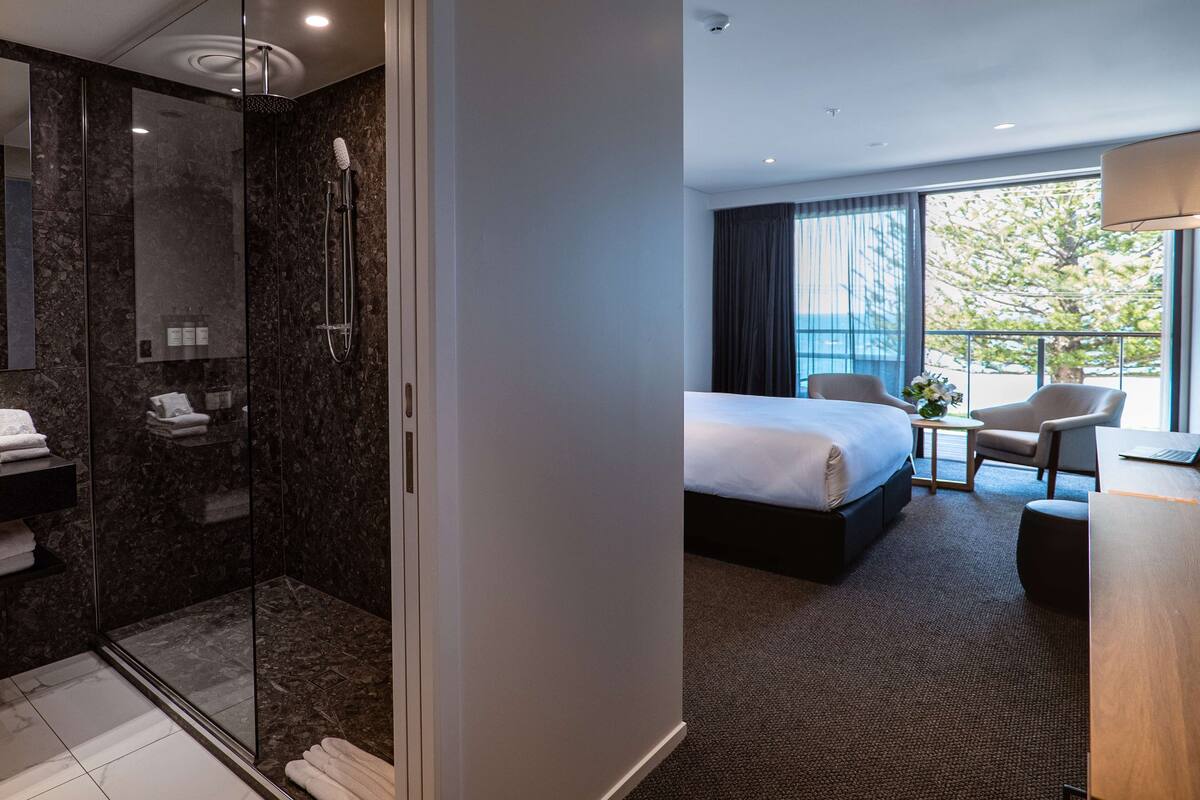 Een moderne hotelkamer met een douchecabine van glas, een groot bed, een bureau en uitzicht op het omliggende landschap.