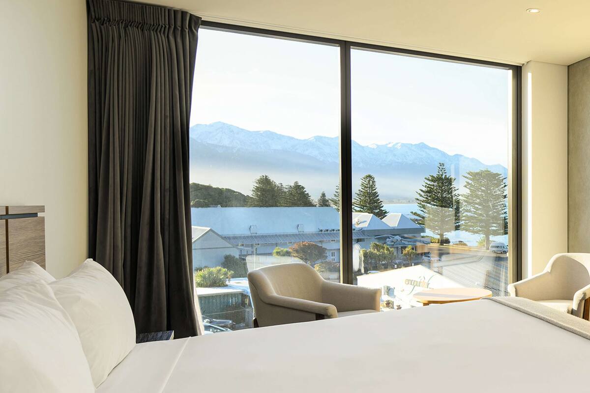 Een hotelkamer met een groot bed, twee stoelen, een klein tafeltje en uitzicht op bergen door grote ramen.