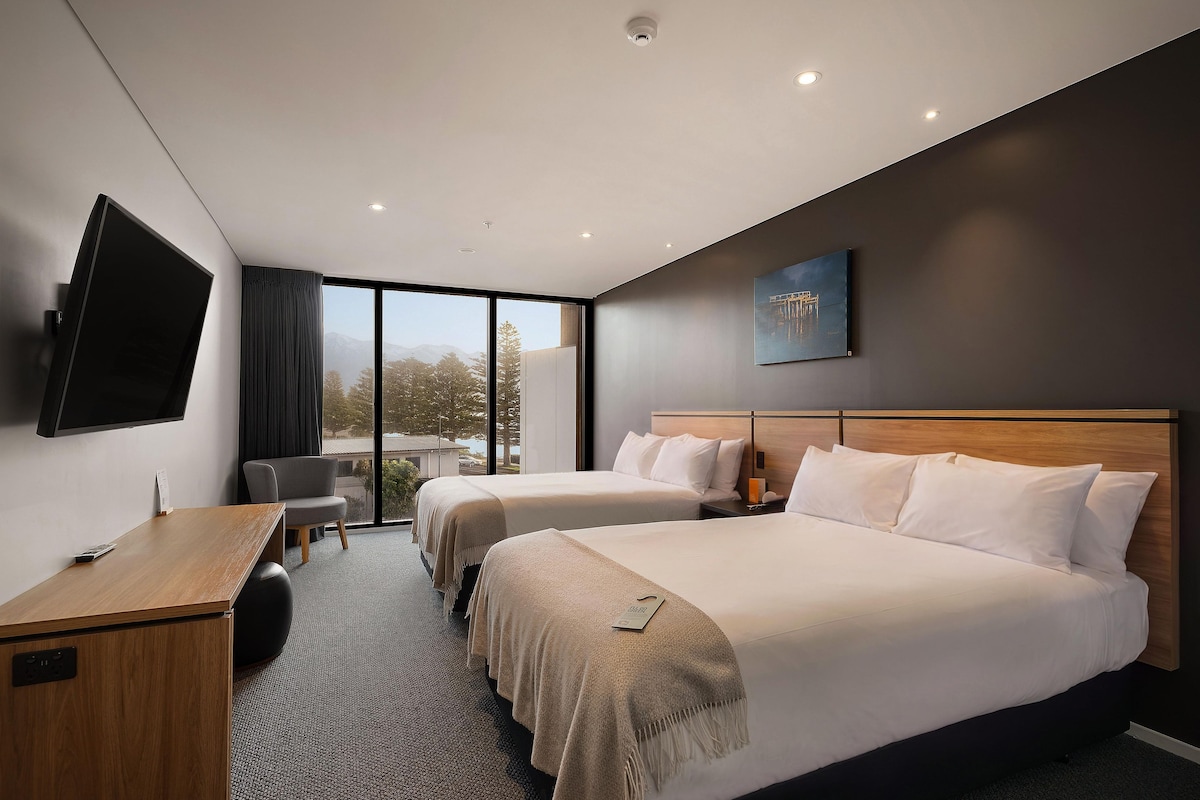 A hotel room with two beds, a desk, a chair, a flat-screen TV, and a painting on the wall.