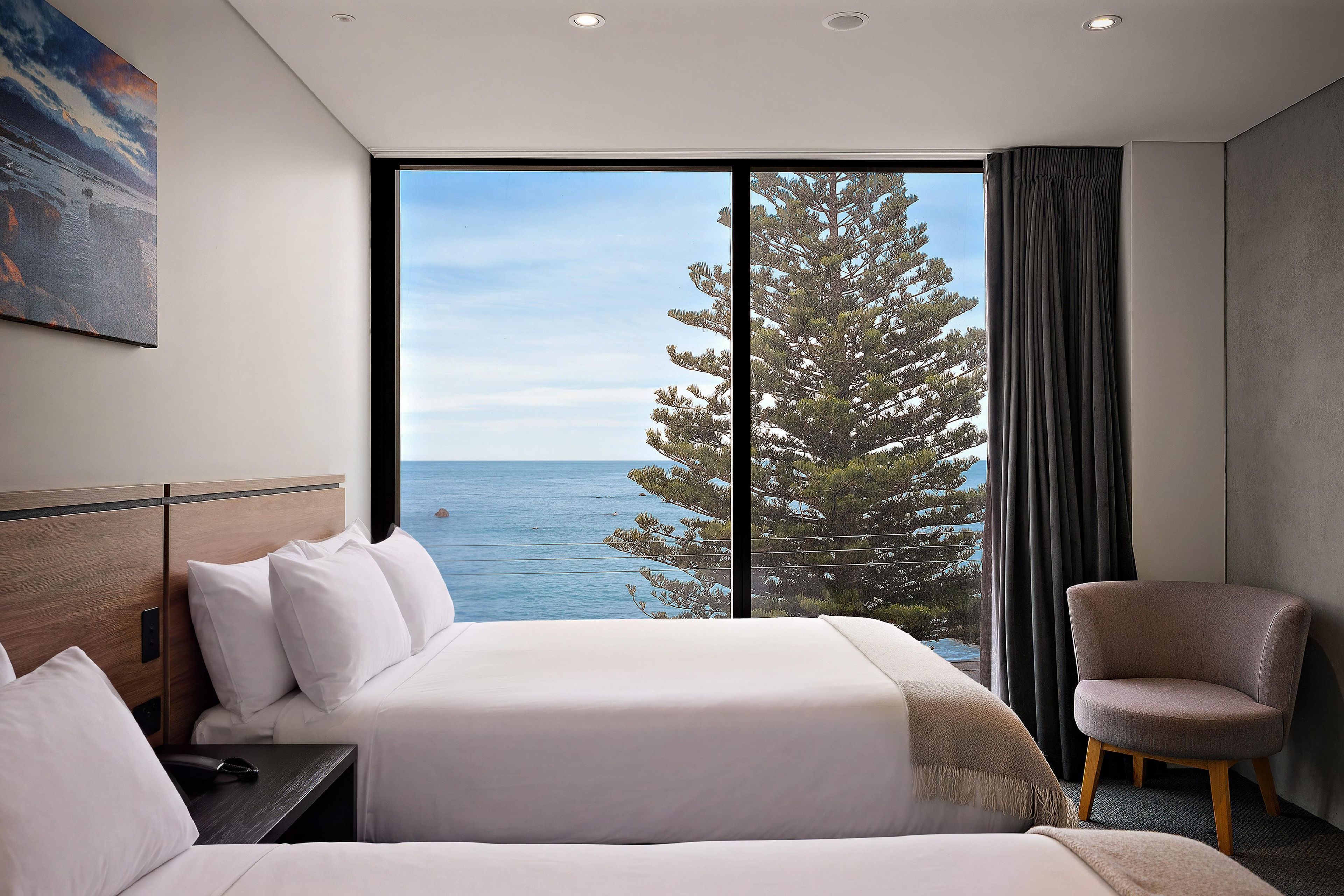 A hotel room with a large window, a bed, a chair, and a painting on the wall.