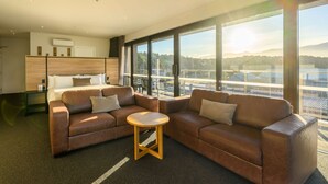 Une chambre d’hôtel moderne avec une grande fenêtre, un canapé en cuir marron, une petite table ronde et un lit doté d’une tête de lit en bois.