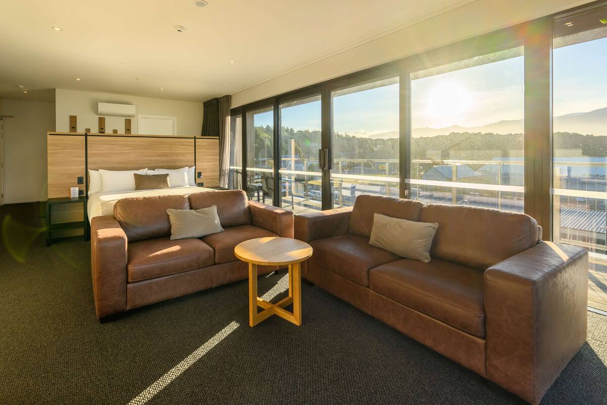 Een moderne hotelkamer met een groot raam, een bruine leren bank, een klein rond tafeltje en een bed met een houten hoofdbord.
