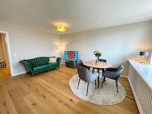 En moderne stue med en grøn sofa, et rundt træbord med stole, et fjernsyn og en dekorativ vægdekoration.